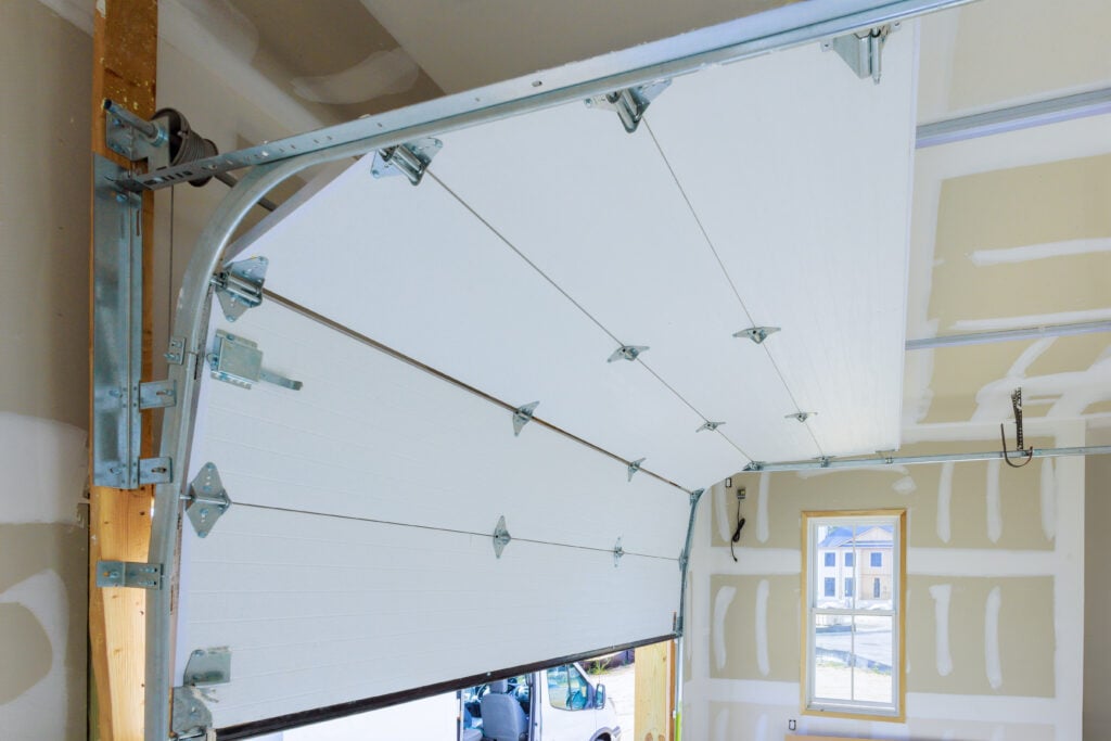 Torsion spring garage door system viewed from inside a garage, showing cables, tracks, rollers, and hinges during installation